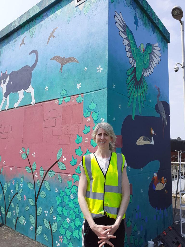 The artist, Caroline Daly, stands in front of her new mural at Chorlton tram station